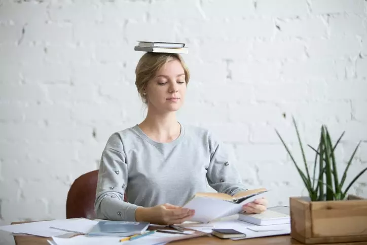 Portrait of attractive woman at desk, books on her head, showing good posture