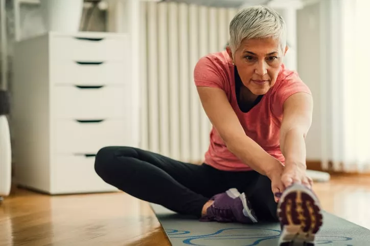 Mature woman exercising at home. Sitting on exercise mat and working extremity stretches on her legs.