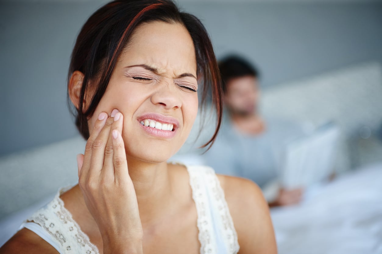 Shot of a woman sitting on the side of her bed with jaw pain with her boyfriend in the background. may be experiencing a condition known as Bruxism.