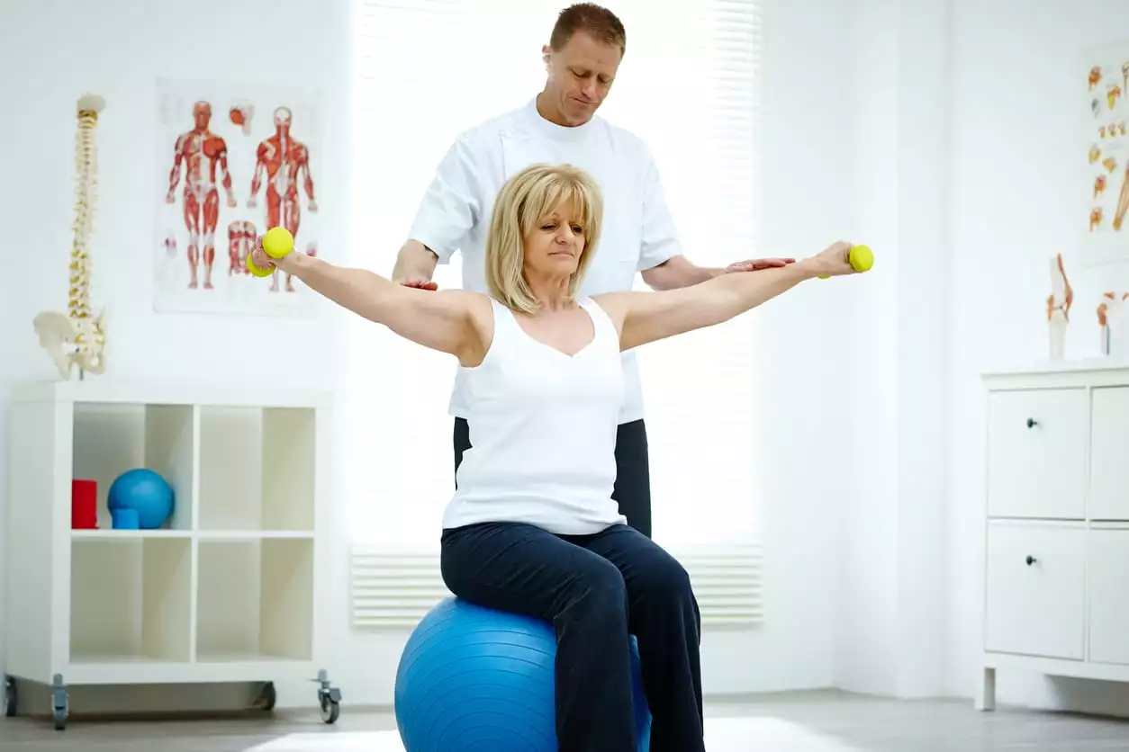 Senior woman sitting on fitness ball holding dumbbells being assisted by her physiotherapist in medical room. Rotator cuff injury concept