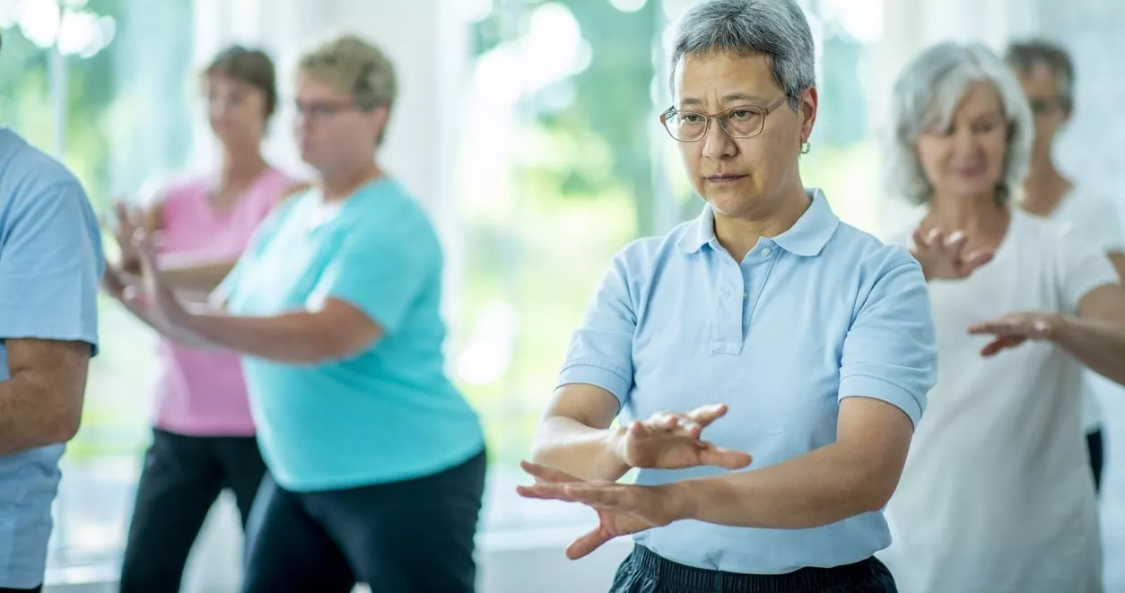 A group of senior individuals are indoors. They are performing the tai chi technique of meditation to attain mental and physical wellbeing.