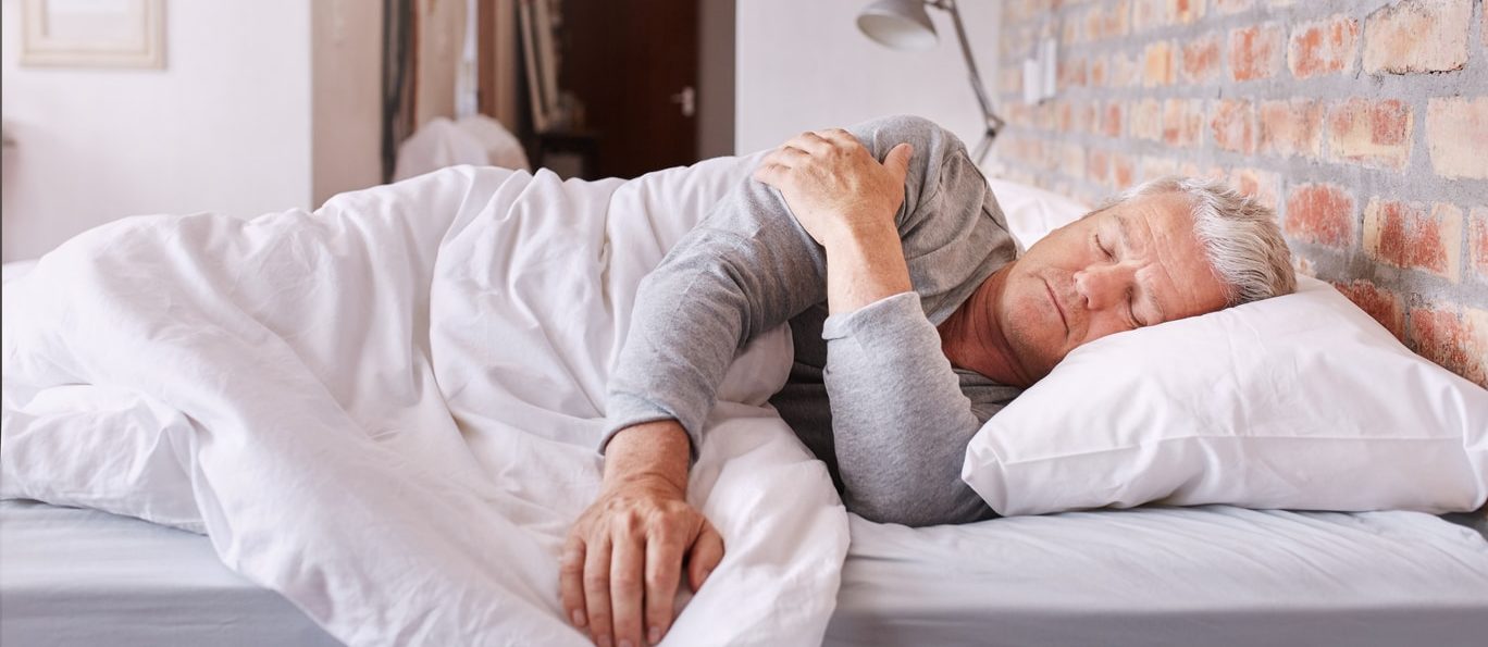 Shot of a senior man sleeping in bed holding his shoulder because of shoulder pain at night