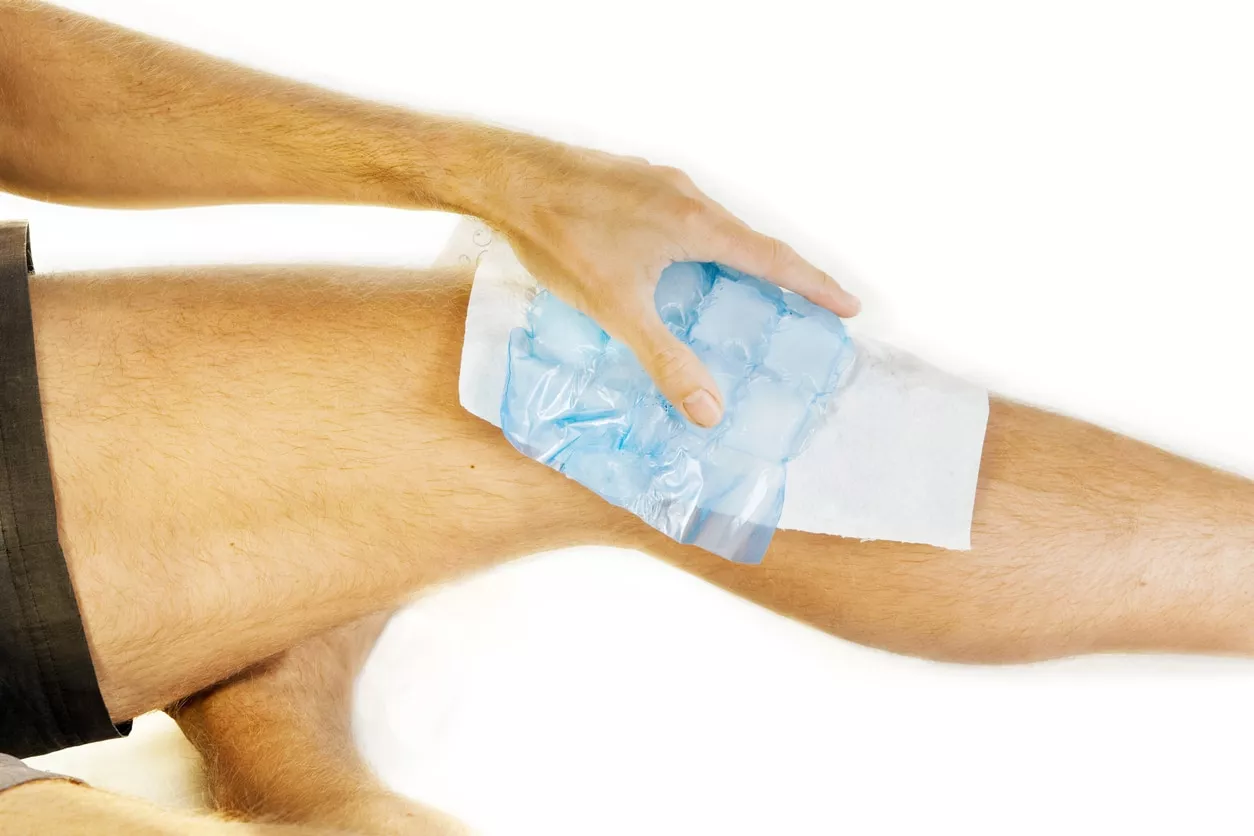 Lots of ice on an aching knee joint. A sheet is placed between ice and skin. A hand is holding the ice. XXL size image. RICE method
